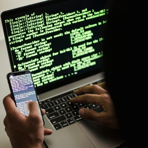 A hooded figure engaged in hacking using a laptop and smartphone in low light.